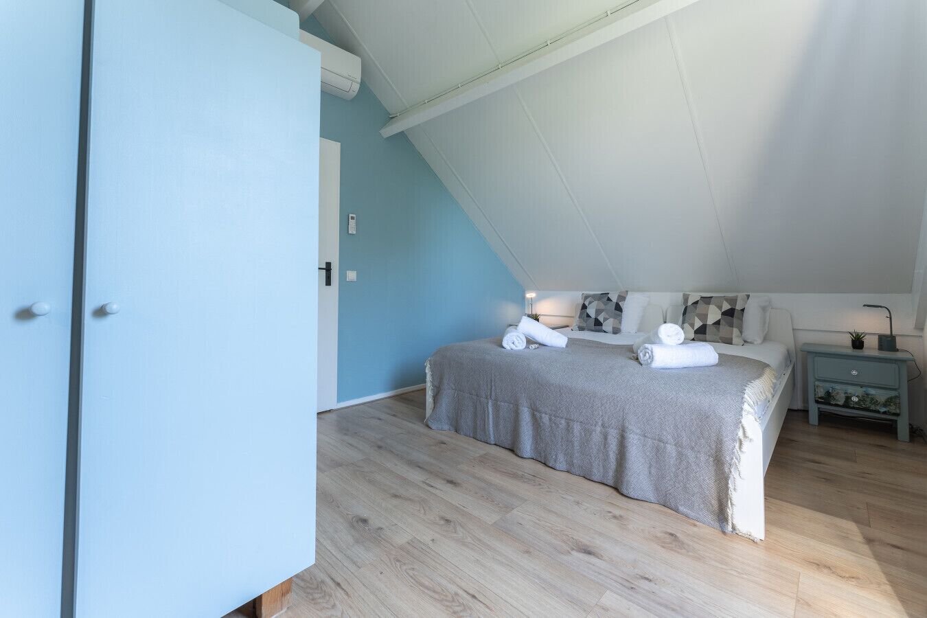 Une chambre avec un plafond en pente, un lit recouvert d’une couverture grise, deux tables de chevet avec des lampes et une armoire.