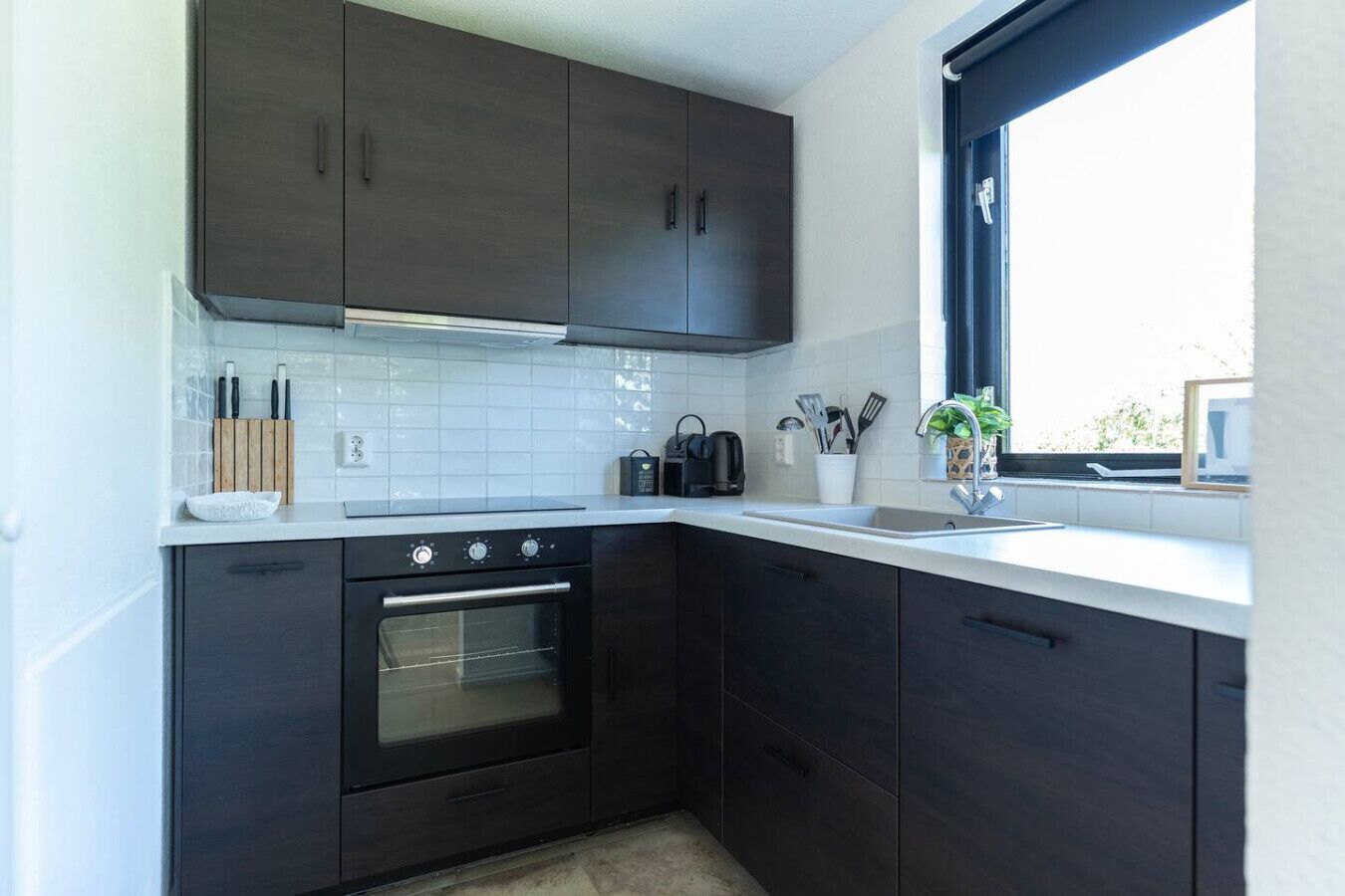 Eine moderne Küche mit dunklen Schränken, einem eingebauten Backofen, einer weißen Arbeitsplatte und einem Fenster mit Aussicht.