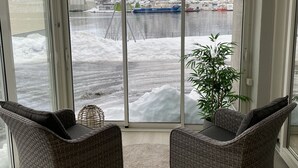 Une pièce avec deux fauteuils en osier, une plante en pot et une vue sur un paysage enneigé avec un navire.