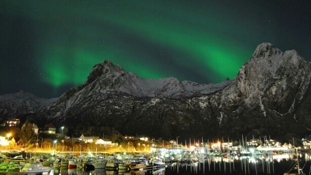 Une vue nocturne d’un port, avec des bateaux, des montagnes enneigées et une ville illuminée par des lumières et bordée de bâtiments.
