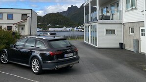 Une voiture noire est garée dans une allée, avec un bâtiment blanc et des montagnes en arrière-plan.