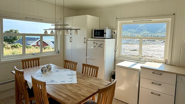 Un coin repas avec une table et des chaises en bois, une cuisine équipée d’un four à micro-ondes et d’un réfrigérateur, et de grandes fenêtres offrant une vue sur la mer.
