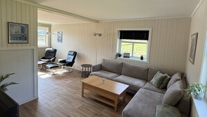 Un salon spacieux avec un grand canapé, une table basse et un téléviseur. Il y a deux fauteuils et une petite table. Le sol est en bois et la pièce est dotée d’une fenêtre avec des stores.