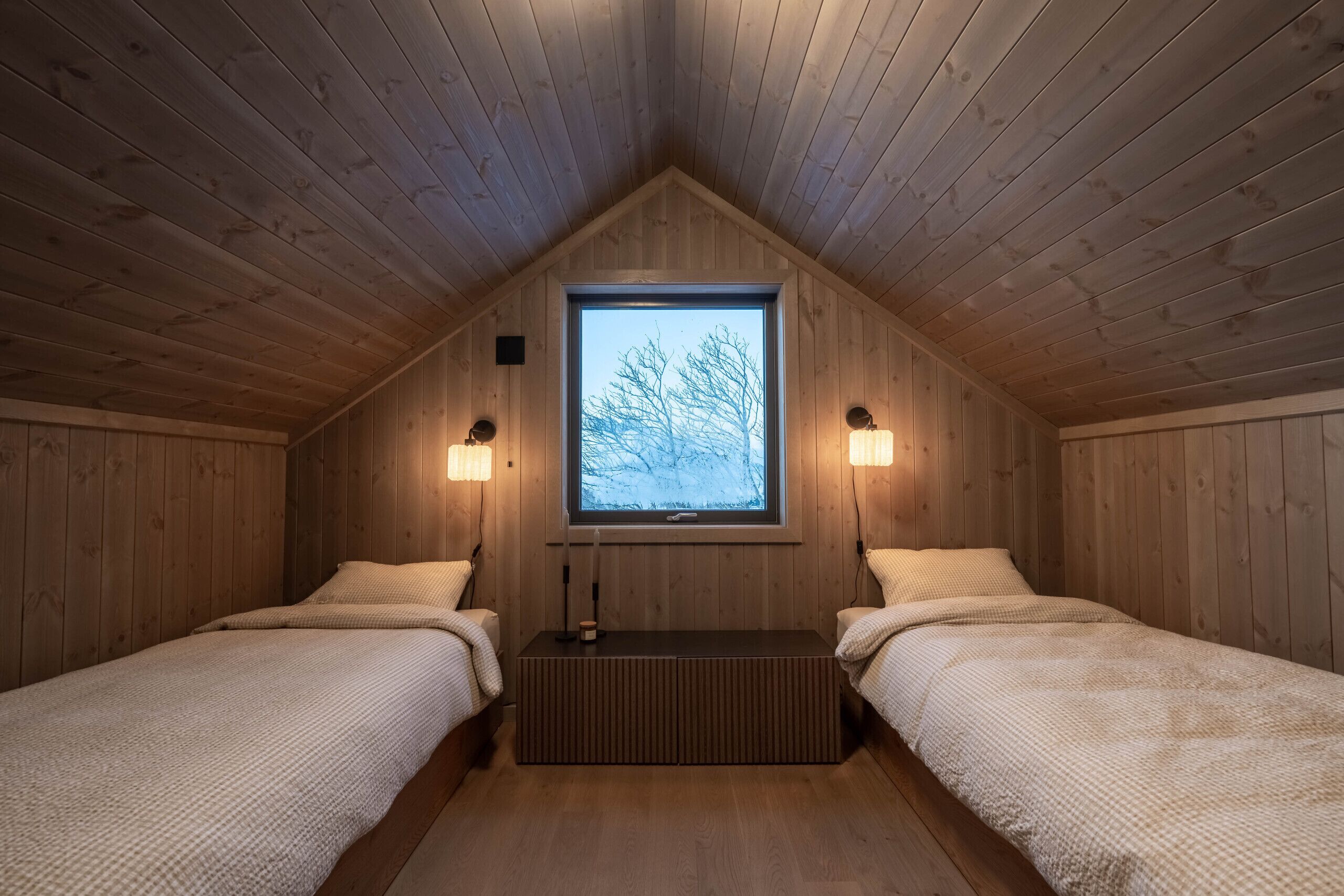 Ein gemütliches Holzhaus mit zwei Betten, einem Tisch in der Mitte und einem Fenster mit Blick auf Bäume.