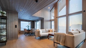 A spacious room with a wooden ceiling, large windows, a sofa, a dining table, and a cabinet with dishes.