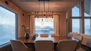 A wooden interior with a dining table, chairs, a vase with flowers, and large windows with a snowy landscape view.