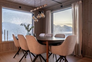 Een eetgedeelte met een houten tafel en stoelen, grote ramen met uitzicht op besneeuwde bergen en hangende lampen.