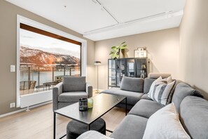 Ein modernes Wohnzimmer mit einer Couch, Sesseln, einem Couchtisch, einem Schrank und einem großen Fenster mit Blick auf ein Gewässer und Berge.