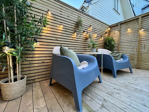 Eine Holzterrasse mit zwei blauen Stühlen, einem Blumentopf und Lichterketten.