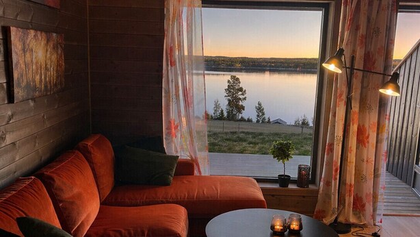 A cozy room with a wooden interior, a red sofa, a small round table with candles, and a view of a lake. - Modern apartment in Golsfjellet (Gol)