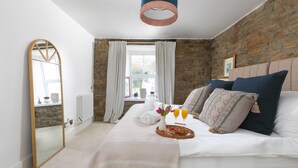 Ein Schlafzimmer mit Steinwand, einem Bett mit weißer Bettwäsche, einem Spiegel, einem Fenster mit Vorhängen und einem Frühstücksbrett mit Obst und Saft.
