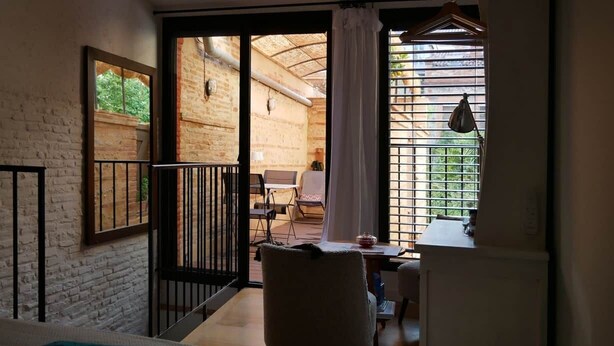 Ein Schlafzimmer mit Bett, Schreibtisch, Stuhl und Fenster mit Blick auf eine Ziegelwand und Grünpflanzen.