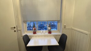 Une pièce d’angle avec une porte blanche, une fenêtre avec des stores, deux chaises, une petite table et un rebord de fenêtre orné de deux statuettes.