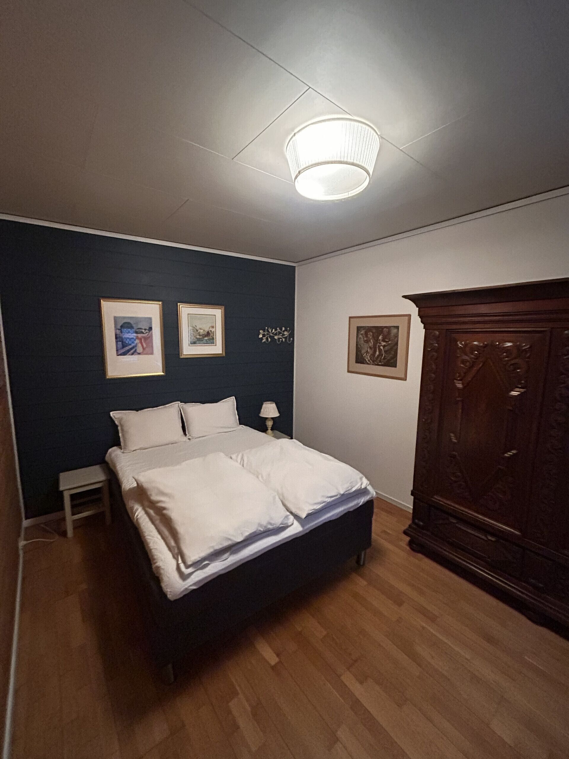 Een slaapkamer met een bed, een nachtkastje, een dressoir en lijstjes met kunstwerken aan de muur.