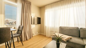 A modern living room with a sofa, a dining table with chairs, a flat-screen TV mounted on the wall, and a window with curtains. - Lofoten Premium Suites #Aurora (Vestvågøy)