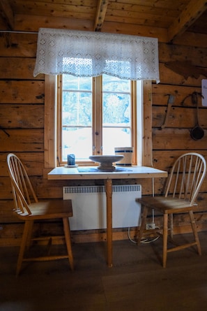 Ein Holzhütten-Interieur mit Tisch, zwei Stühlen, einem Fenster mit Spitzenvorhang und einem weißen Heizkörper.