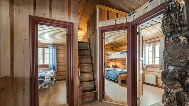 Un intérieur en bois avec un escalier menant à une chambre en mezzanine, un mur en pierre et une vue dégagée sur l’extérieur par une porte ouverte.