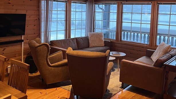 A cozy cabin interior with wooden walls, a sofa, chairs, a TV, and large windows offering a view of the outdoors. - Pollstua, cosy cabin with amazing views (Vestvågøy)