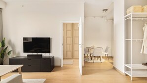 Ein modernes Wohnzimmer mit Flachbildfernseher, einem dunklen Holzschrank, einem Essbereich mit Tisch und Stühlen sowie einem weißen Kleiderschrank.