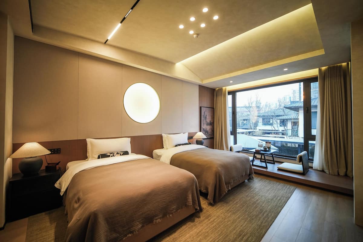 Une chambre d’hôtel avec deux lits, une grande fenêtre donnant sur un beau paysage, un applique rond au mur et une petite table.