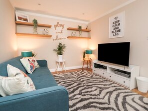A living room with a blue sofa, a flat-screen TV, wooden shelves with plants, a wall-mounted sign, and a patterned rug. - Bronte View Cottage, Haworth (Keighley)