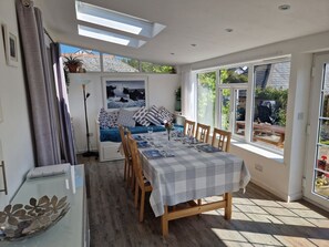 A dining area with a table set for four, a sofa with cushions, and a view of the outdoors through large windows. - Langurra in Crantock, Cornwall (Newquay)