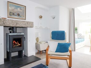 A room with a fireplace, a chair with a blue towel, a painting on the wall, and a view to the outside. - Langurra in Crantock, Cornwall (Newquay)