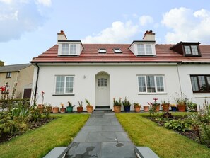 A two-story house with a red-tiled roof, a central entrance, and a garden with potted plants.