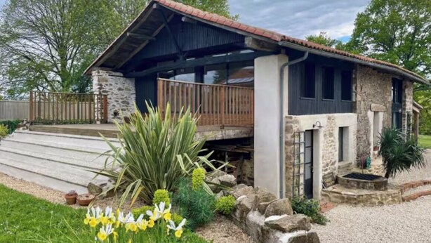Un bâtiment rustique en pierre, doté d’une terrasse en bois, entouré d’une allée en gravier et d’un jardin où poussent des fleurs jaunes.