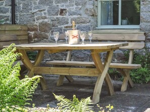A wooden picnic table with a bottle of champagne and glasses, set against a stone wall with a window.