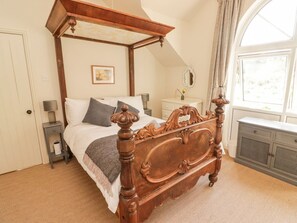 A four-poster bed with a wooden frame, a bedside table with a lamp, a dresser with a mirror, and a window with curtains.