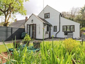 Una casa blanca con techo oscuro, una chimenea visible y un jardín con plantas verdes y flores amarillas.
