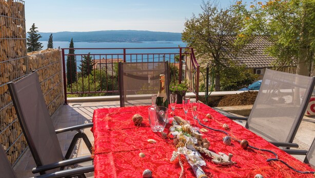Mesa al aire libre con vistas a un lago y montañas, sillas de metal y mantel rojo.