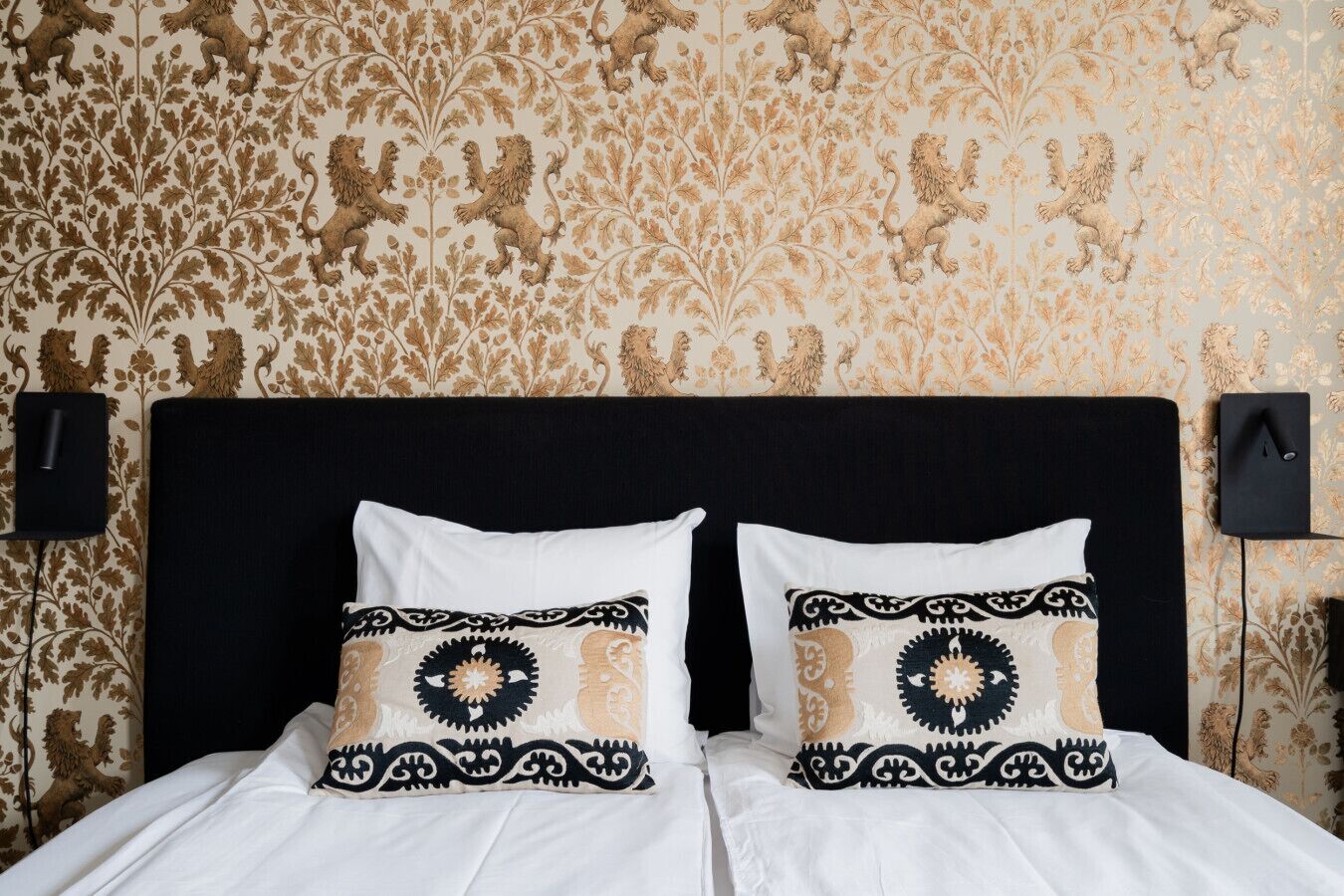 Una cama con dos almohadas, cabecera negra y papel tapiz estampado.
