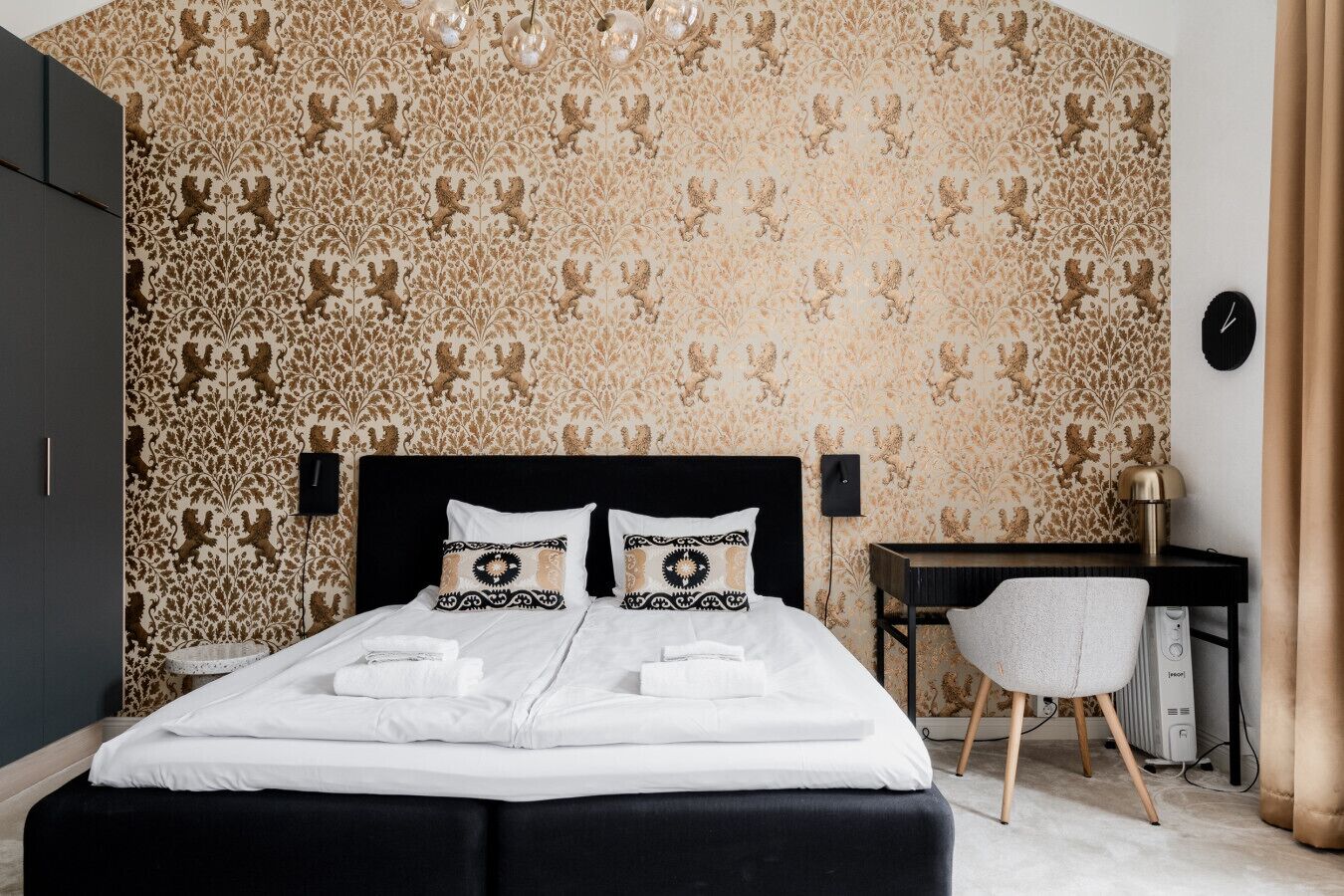 Un dormitorio con papel tapiz estampado, cabecera negra, dos almohadas, mesita de noche, una silla y un escritorio.