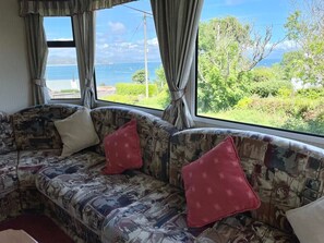 Un sofá con tapicería estampada y cojines rojos, frente a una ventana con vistas a vegetación y un cuerpo de agua.
