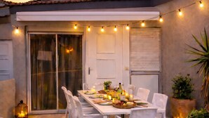 An outdoor dining area with a table set for four, surrounded by white chairs, under string lights. - Air-conditioned house for 6 with supervised swimming pool - Beach & port on foot - Terrace (Saint-Cyprien)