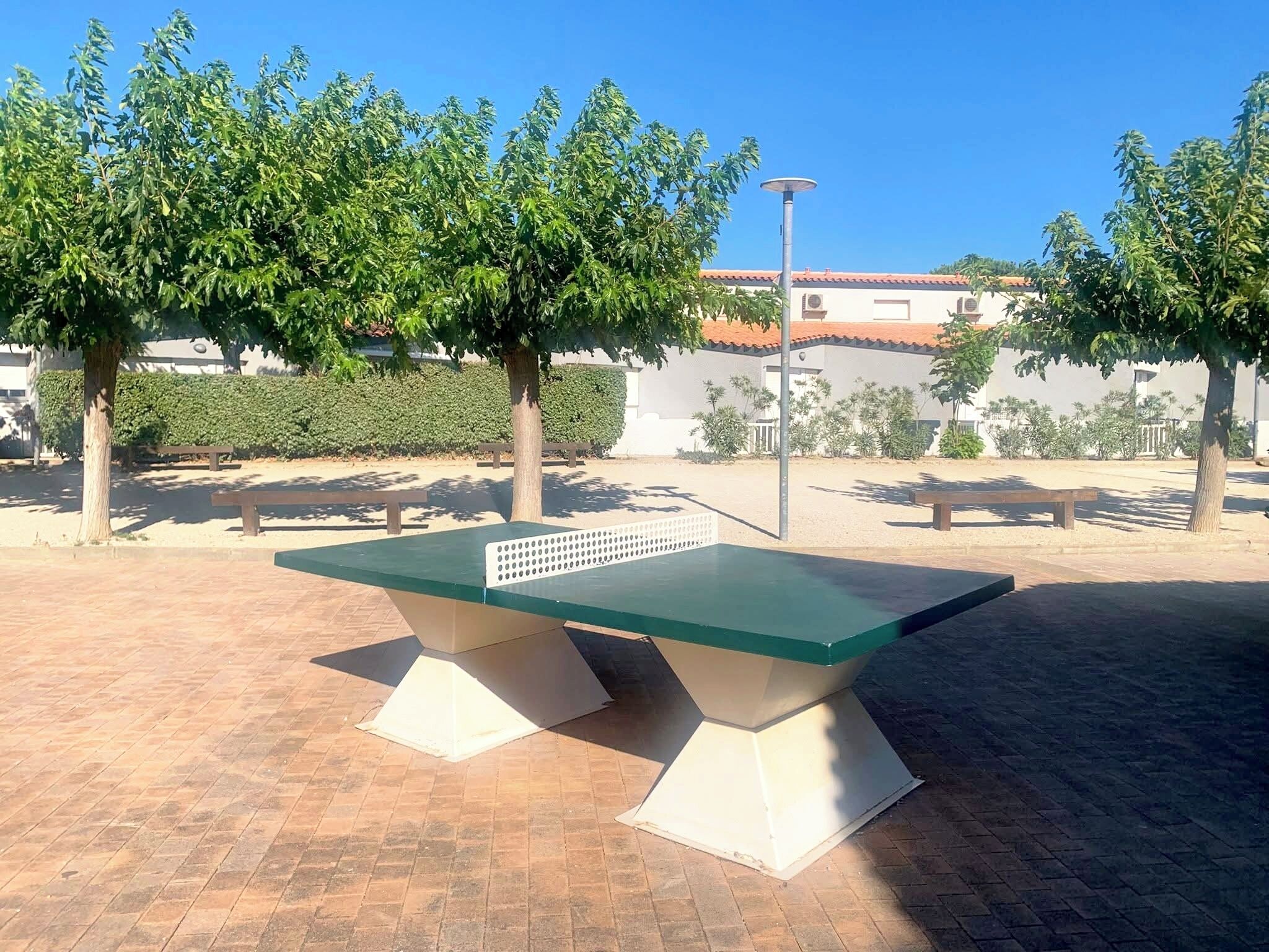 Une table de ping-pong installée dans un espace extérieur, avec des arbres et un bâtiment en arrière-plan.
