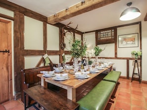 Un comedor con una larga mesa de madera dispuesta para una comida, bancos y un corpulento ciervo colgado.