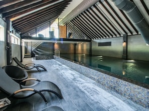 An indoor swimming pool with a tiled floor, black lounge chairs, and a staircase leading to an upper level.