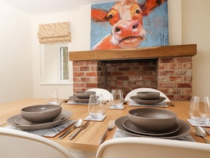 Un coin repas agrémenté d’une cheminée en briques, d’un tableau représentant une vache et d’une table mise pour quatre personnes.