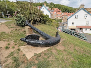 A coastal town with a prominent anchor sculpture on a grassy hill, traditional houses with red roofs, and a paved road.
