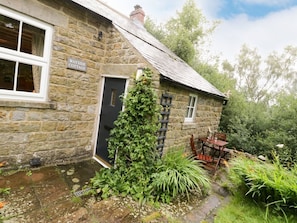 Une petite maison en pierre avec une pancarte indiquant « Wayside Cottage », une porte, une fenêtre, un petit jardin avec des plantes et une table avec des chaises à l’extérieur.