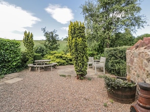 A garden with a gravel patio, a wooden table, and chairs, surrounded by various shrubs and trees. - Near Bank Cottage, St Abbs (Eyemouth)