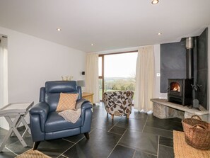A living room with a fireplace, a leather sofa, a patterned armchair, and a view through a large window. - Sleepy Fox, Blisland (Bodmin)