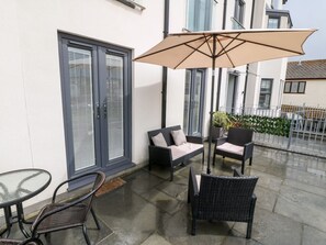 Ein überdachter Außenbereich mit Korbmöbeln und einem Tisch, neben einem Gebäude mit großen Fenstern und einer Haustür mit Fensterläden.