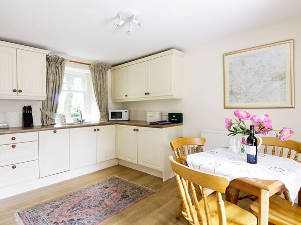 A kitchen with white cabinets, a microwave, a sink, and a dining table with chairs. There is a window with curtains, a vase with flowers, and a framed picture on the wall.