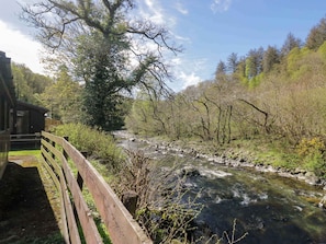 A wooden bridge over a flowing river, surrounded by trees and a building on the left.