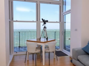 A room with a table and chairs, a telescope on a tripod, and a balcony with a railing. - 4 Oceanside Apartments, Ilfracombe (Ilfracombe)