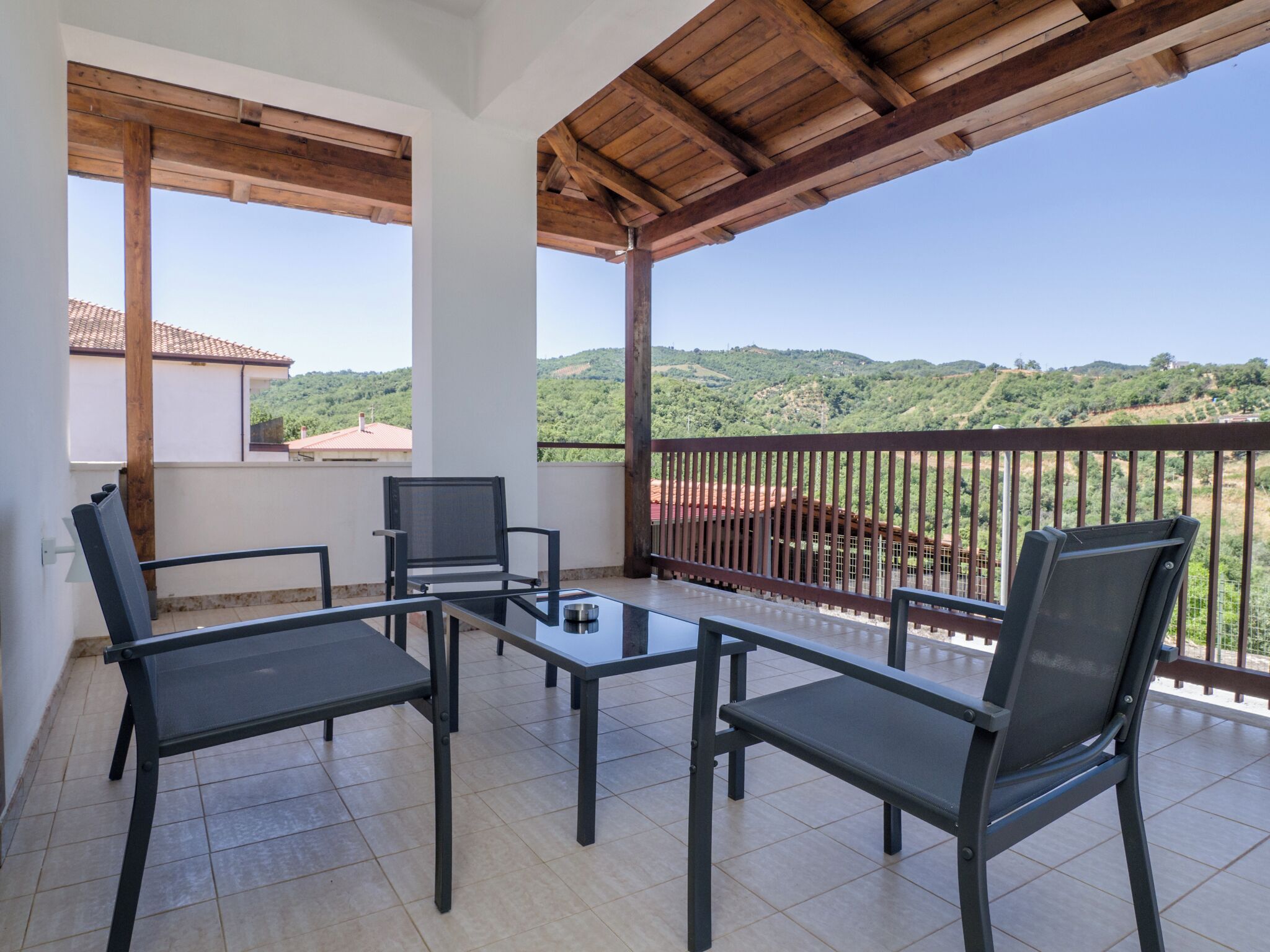 A balcony with a table and chairs, a view of a hilly landscape, and a wooden ceiling.